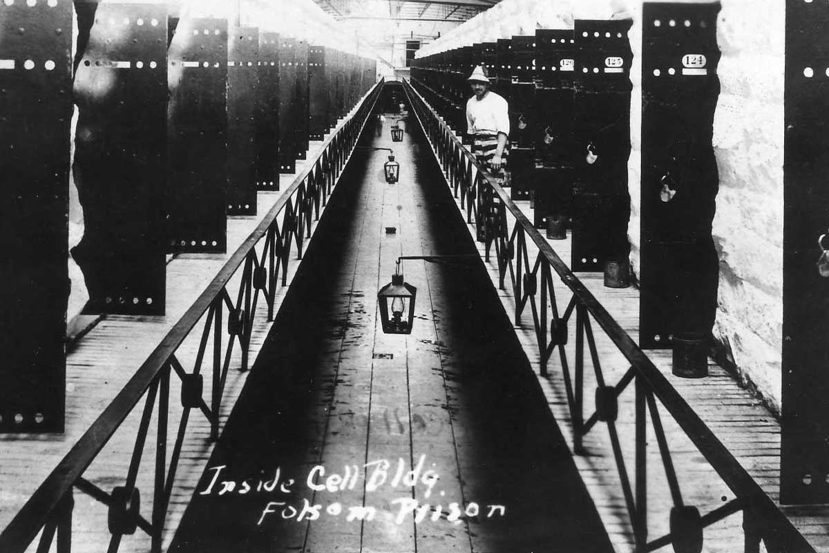 A historical black-and-white photograph showing the interior of a cell block at Folsom Prison. The image captures a long corridor flanked by rows of cells on either side. A prisoner in striped clothing stands near one of the cells. The corridor features overhead lanterns and a handwritten label at the bottom reads, 'Inside Cell Bldg, Folsom Prison.'