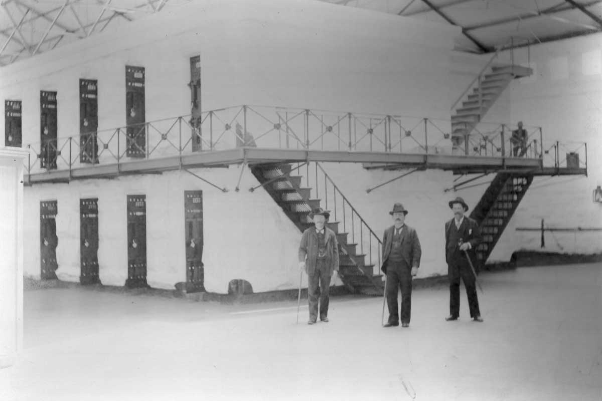 A historical black-and-white photograph of an interior cell block at Folsom Prison. The image shows a two-tiered cell block with metal staircases and walkways. Three men in suits, likely prison staff, stand in the foreground, while a prisoner can be seen seated on the upper level. The cell doors are lined up in rows on both levels, with the structure enclosed by a high ceiling.