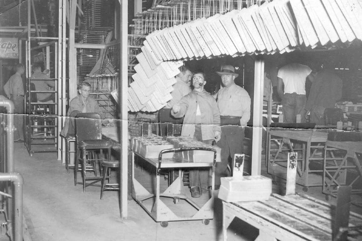 A historical photograph of the license plate manufacturing area at Folsom Prison. The image shows several inmates working on the production line, assembling and painting license plates for California. The workspace is filled with machinery, workstations, and partially completed plates hanging overhead. Prison staff can be seen supervising the process, ensuring the operation runs smoothly.