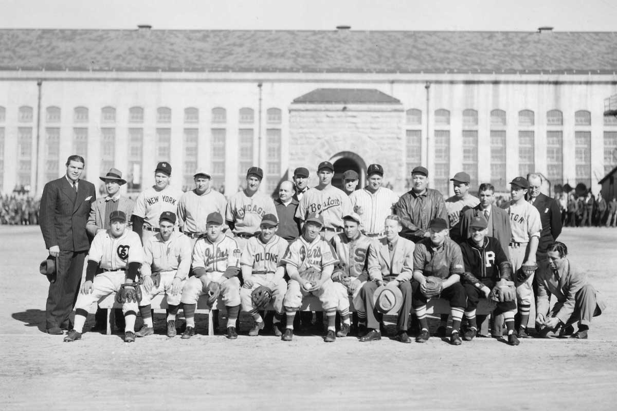 A historical photograph of the Folsom Prison baseball team. The image shows the team members in their uniforms posing for a group photo in front of the new cell building. The players are dressed in various baseball uniforms, some with recognizable team names like New York, Chicago, and Boston. Prison staff and possibly visitors are also seen in the photo.