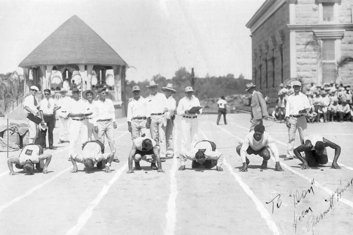 A historical photograph taken at Folsom Prison depicting an outdoor race event. The image shows several inmates in a starting position on a track, ready to begin a race. They appear to be participating in a unique 'peanut race,' where contestants push peanuts along the ground with their noses. Spectators and officials, some in uniforms, stand nearby, overseeing the event. In the background, the bandstand and the chapel are visible.